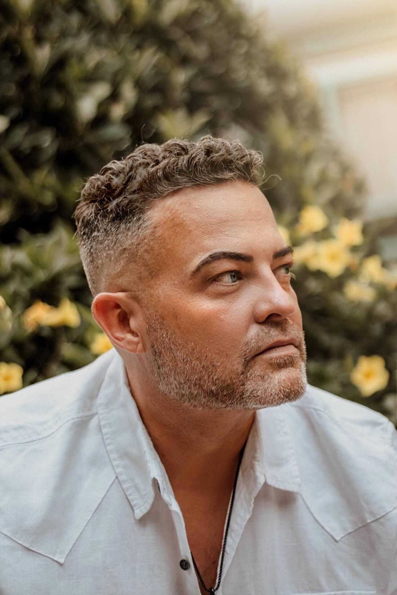 A man with short hair gazing to the right, surrounded by yellow flowers.