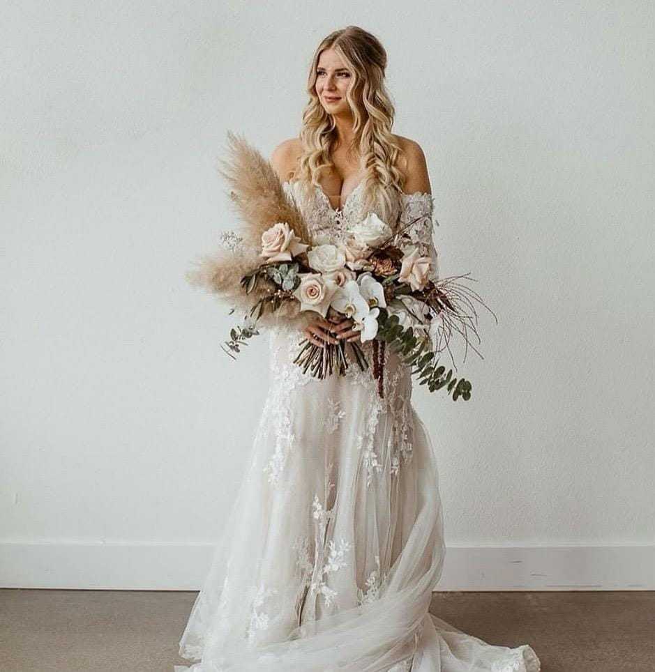 Bride in lace gown holding a bouquet with pampas grass and roses against a plain background.