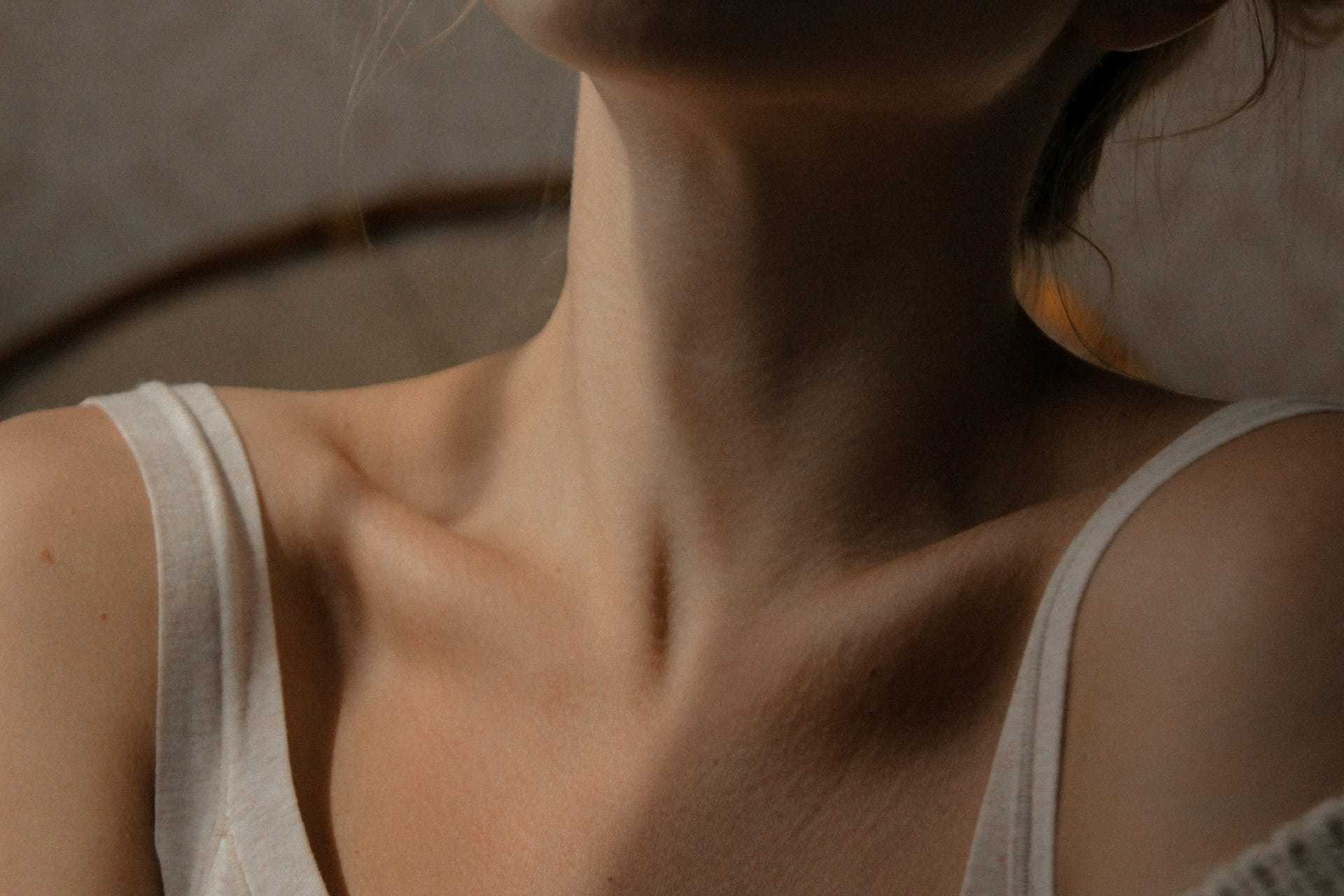 Relaxed woman in a softly-lit room with focus on her neck and shoulders, wearing a white top.