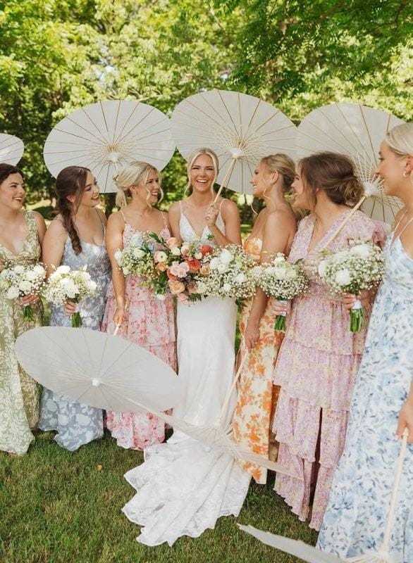 Bridal party with parasols and floral dresses, smiling in a garden setting.